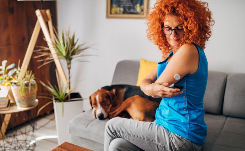 Middle-age red-headed woman checks the glucose monitor on her arm, while sitting on her sofa with a sleep dog lying next to her.