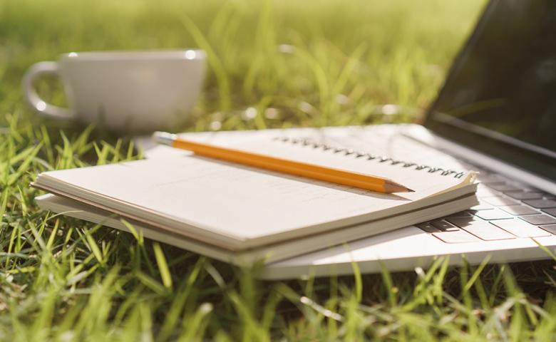 Laptop on grass with notebook and pencil on top and a mug nearby