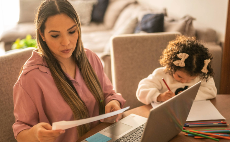 Woman sitting at her kitchen table with a laptop while a small child colours beside her