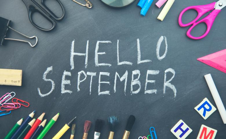 The words &quot;Hello September&quot; written in white chalk on a chalkboard, surrounded by various school supplies.
